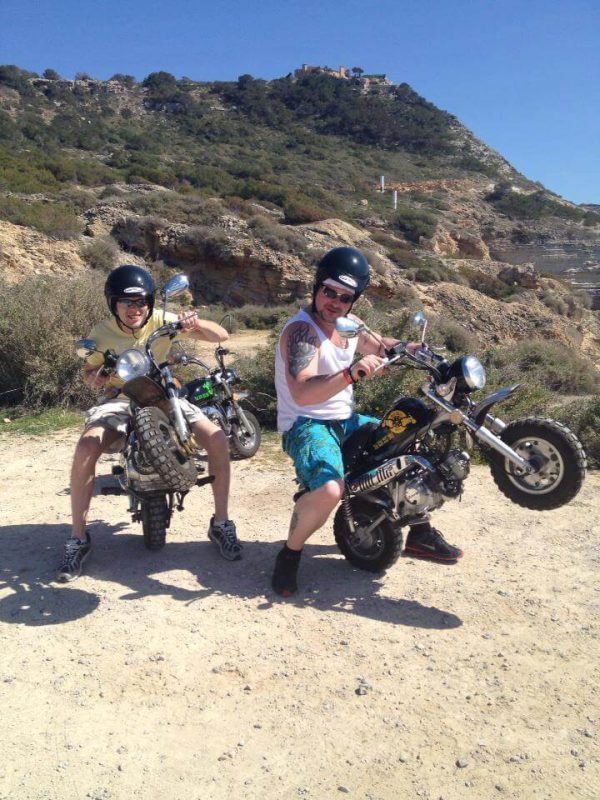 Zwei Männer machen Wheelies auf Minibikes vor mallorquinischer Landschaft, minibike mallorca